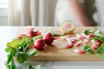 Red radishes on a wood cutting board, with a woman walking by through the kitchen