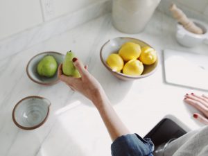 Photo of a woman's left hand holding a green pear. Bowl of lemons and a MAC computer are sitting on the kitchen counter