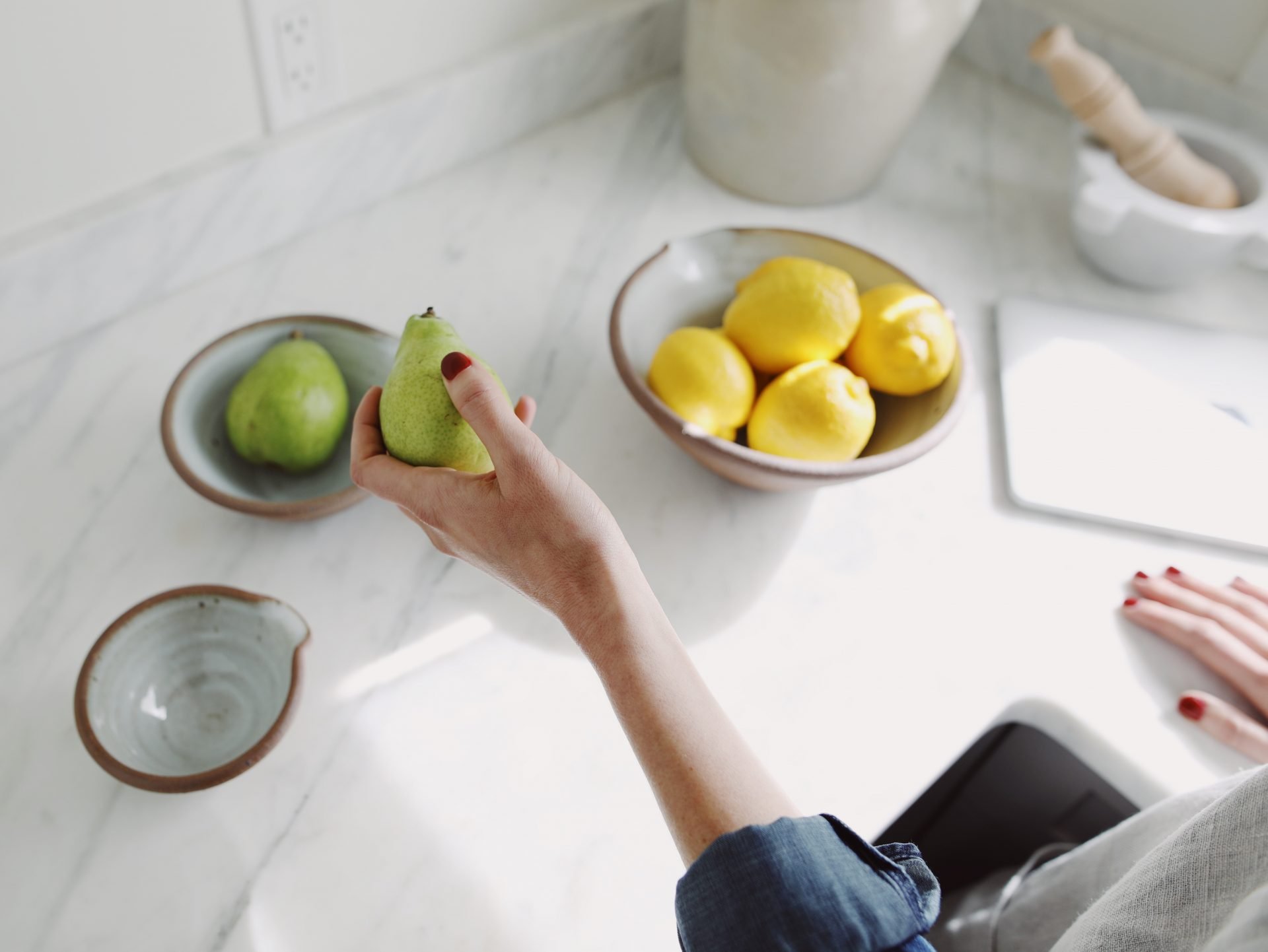 Photo of a woman's left hand holding a green pear. Bowl of lemons and a MAC computer are sitting on the kitchen counter