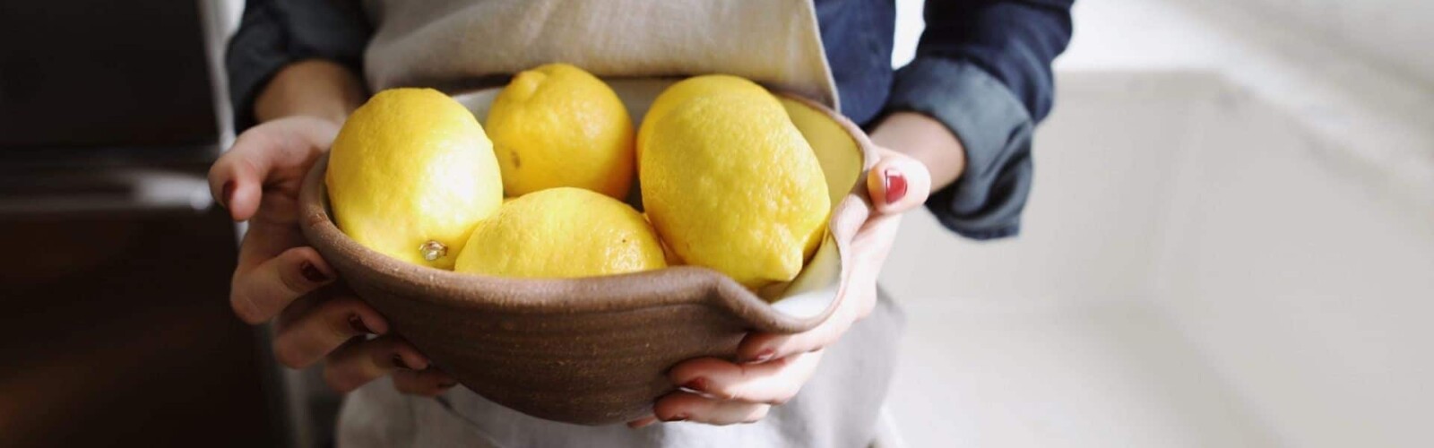 Photo of a woman in an apron holding a bowl full of lemons while standing next to a sink in a kitchen