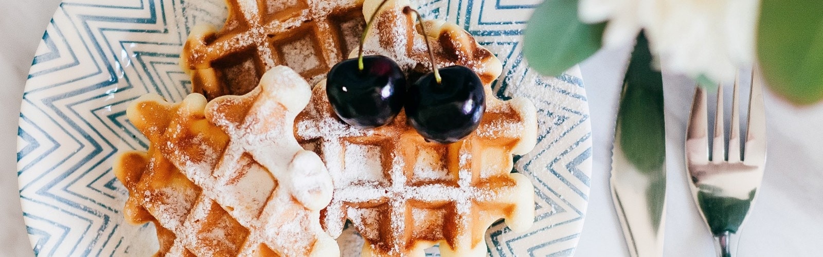 Waffles on a blue plate, topped with red cherries. Fork and knife off to the side